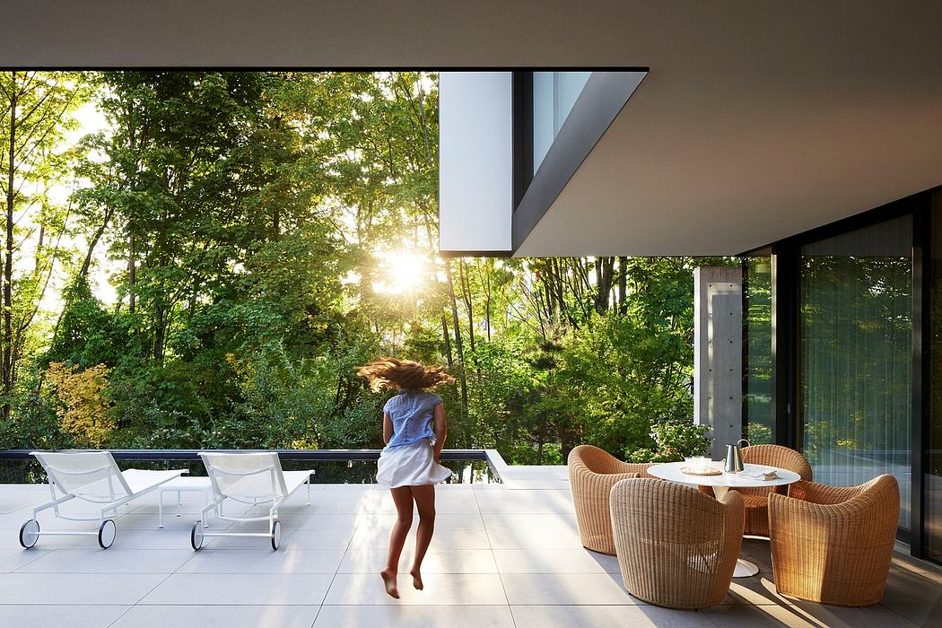 Minimalist patio design with large glass windows overlooking a lush forest, featuring modern seating and dining furniture.