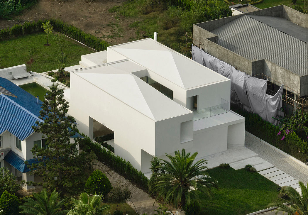 A modern, geometric white house with large windows and a distinctive roof design, surrounded by lush greenery.