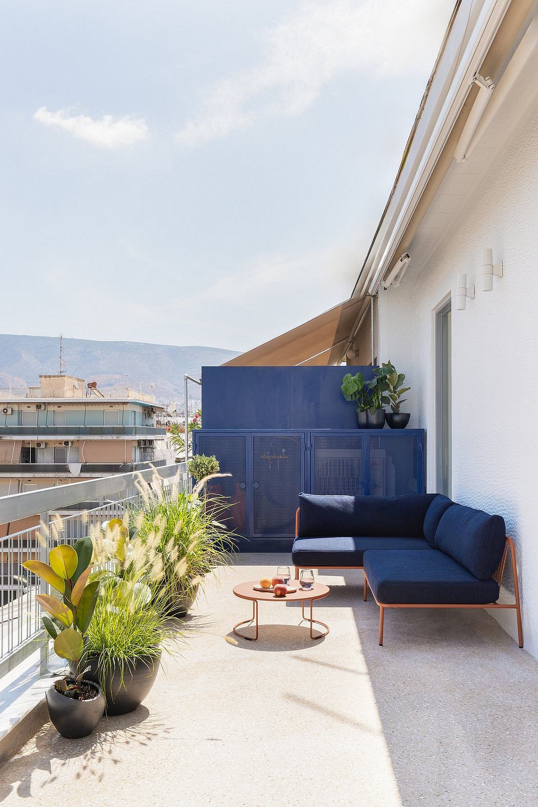 Cozy urban balcony with blue built-in cabinetry, plush seating, and lush potted plants.