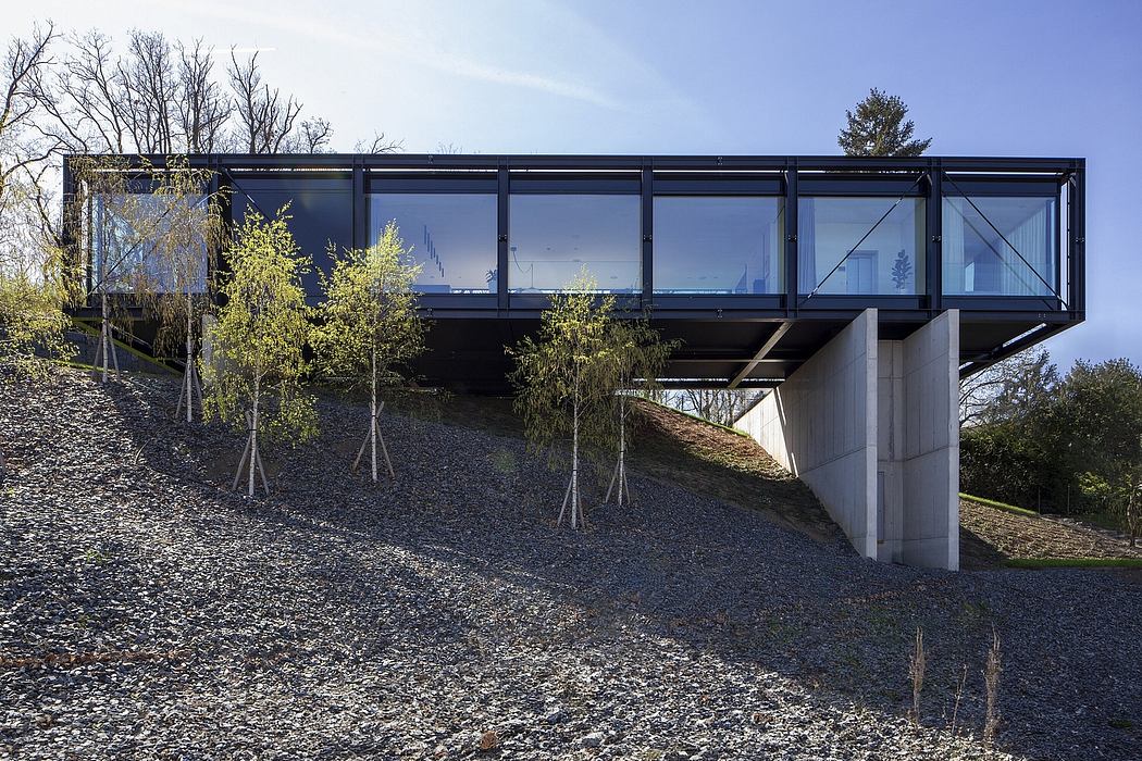 A modern glass-enclosed walkway bridge supported by concrete pillars, surrounded by trees.