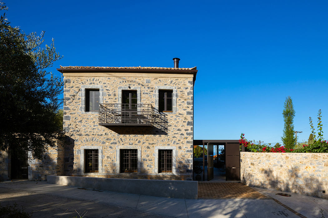 A two-story stone building with a balcony, surrounded by trees and a blue sky.