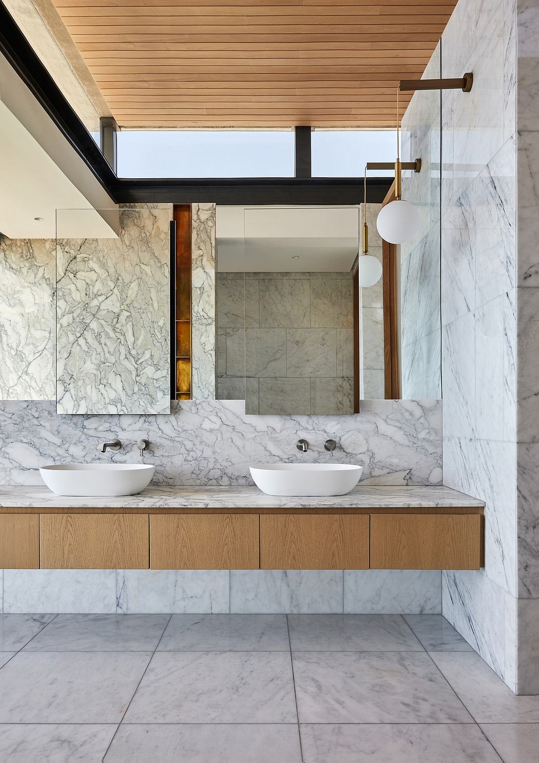 A modern bathroom with marble walls, wooden vanity, vessel sinks, and brass fixtures.