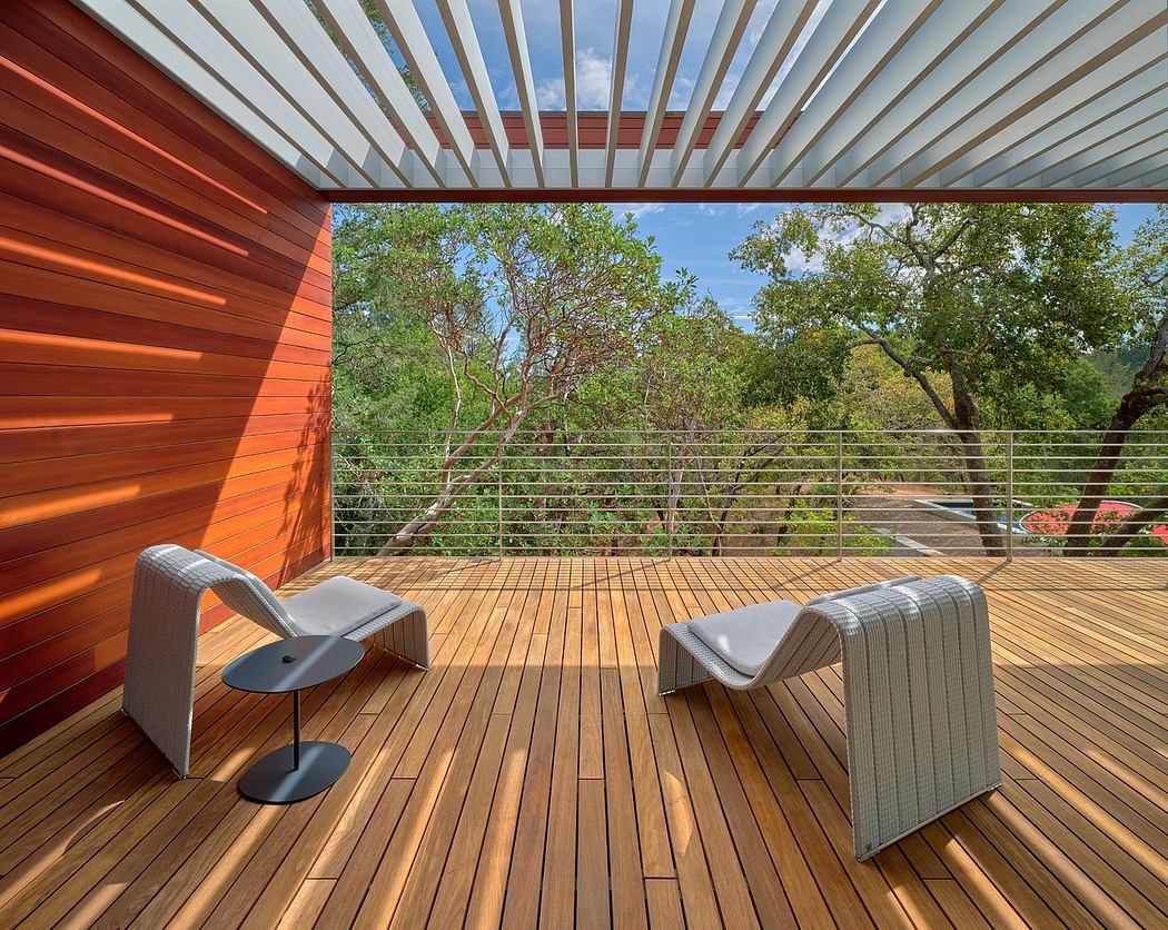 Wooden deck with modern furniture, surrounded by lush greenery and a metal pergola.