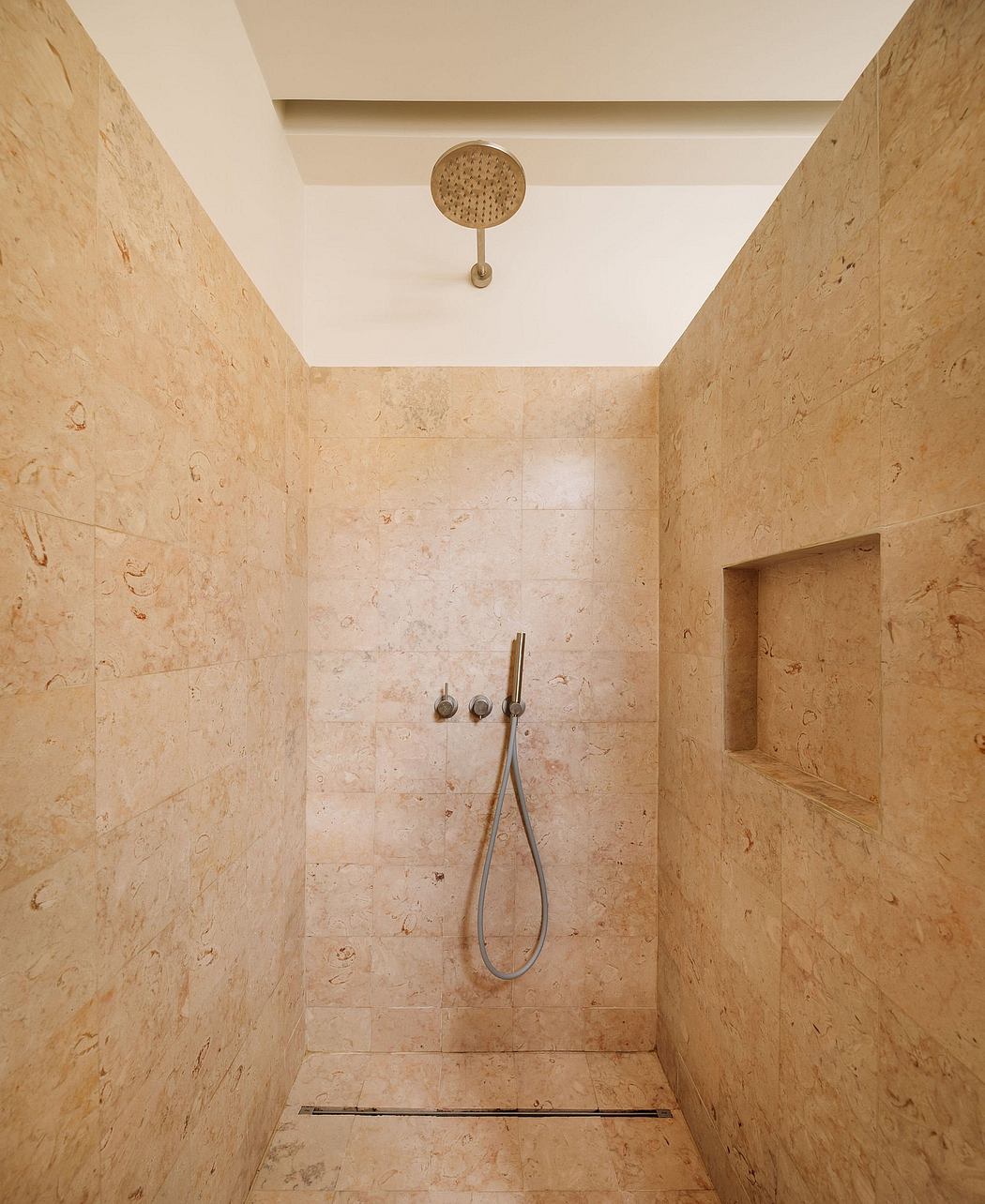 A minimalist shower enclosure with beige natural stone tiles, a round showerhead, and built-in alcoves.