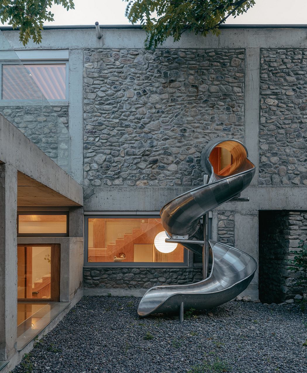 A modern, stone-clad house with a striking metal spiral slide leading to the entrance.