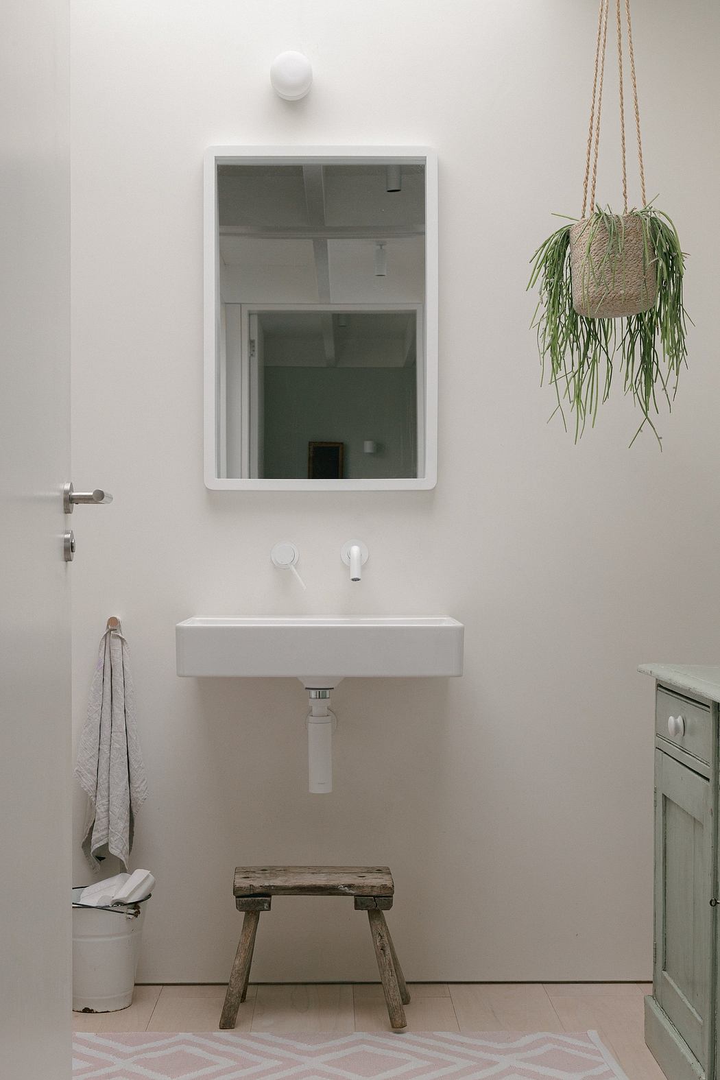 A minimalist bathroom with a white vanity, mirror, and hanging plant, accented by a rustic wooden stool.