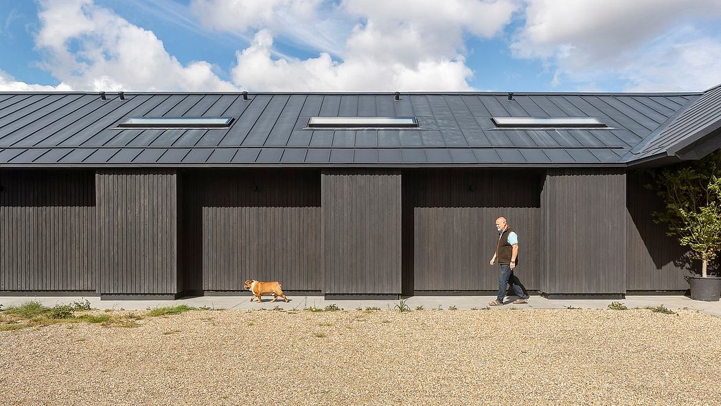 A modern, black-paneled building with large windows and a person walking past a dog on the gravel.