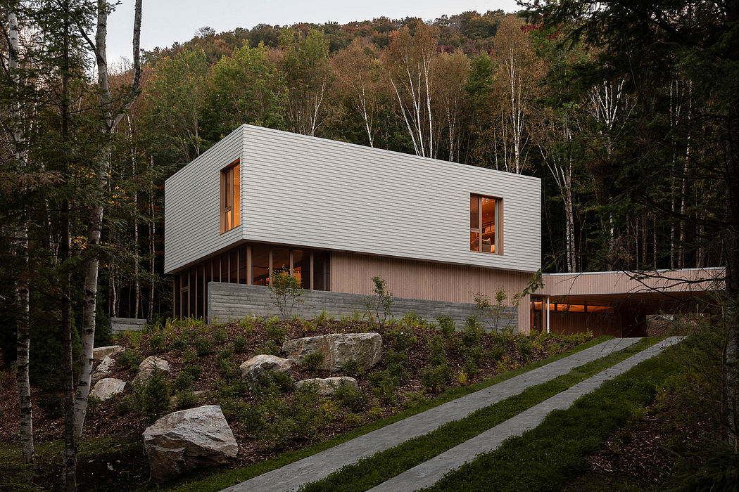 A modern, gray-toned house nestled in a wooded landscape with large windows and a concrete foundation.