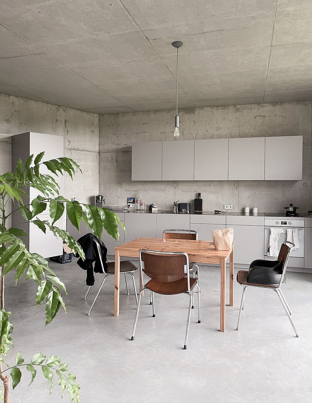 Minimalist kitchen and dining area with concrete walls, white cabinets, and a wooden table.
