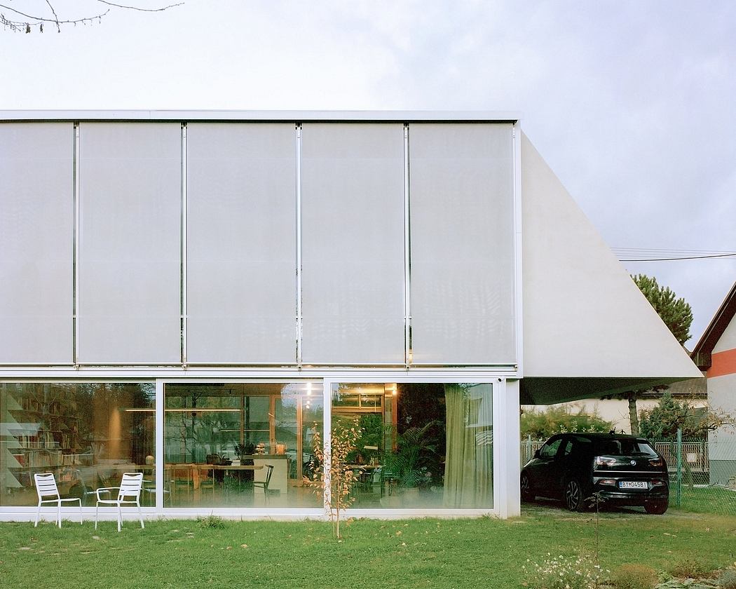 A modern, minimalist glass-walled house with a car parked in the grassy yard.