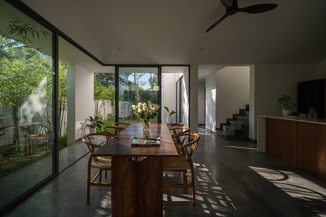 A modern dining room with a large wooden table, comfortable chairs, and lush greenery visible through the glass walls.