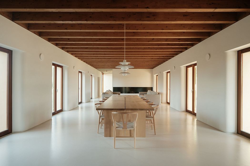 A spacious, minimalist interior with a long wooden table, chairs, and exposed beams on the ceiling.