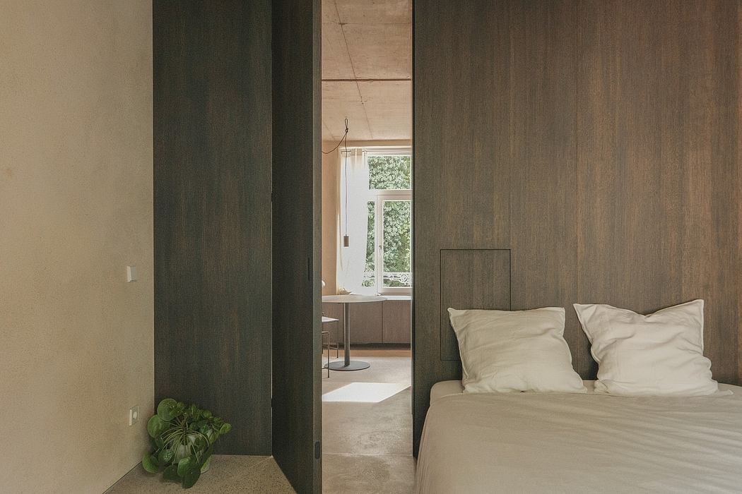 Minimal, warm-toned bedroom with sliding wooden panels, plush bedding, and a potted plant.