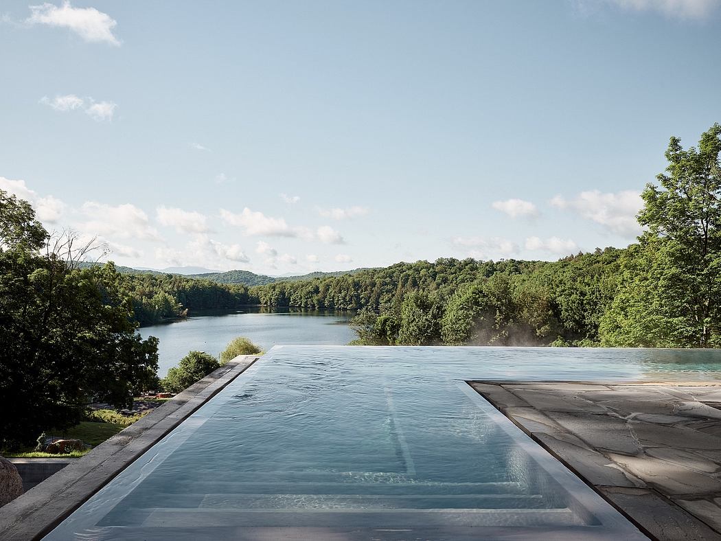 A serene infinity pool overlooking a lush, forested lake against a blue sky.