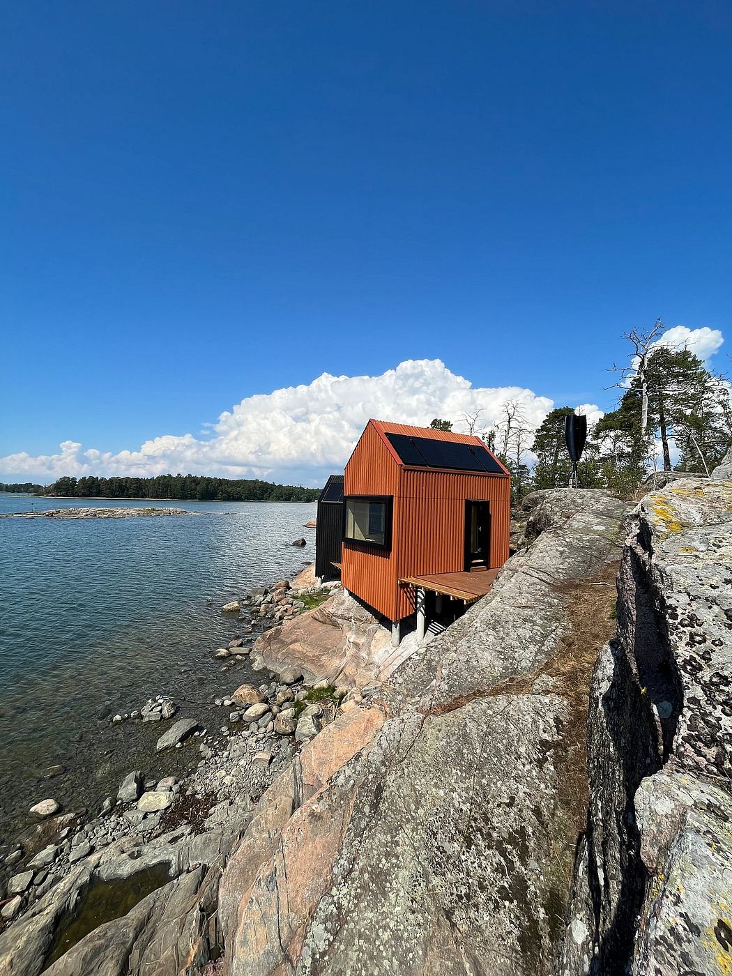 A rustic, red-roofed cabin perched atop a rocky shoreline, overlooking a scenic lake and forest.