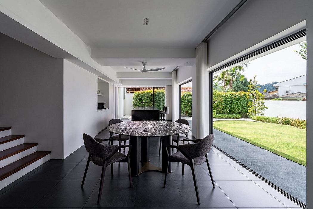 A modern dining area with a round table, sleek black chairs, and floor-to-ceiling windows overlooking a lush outdoor garden.