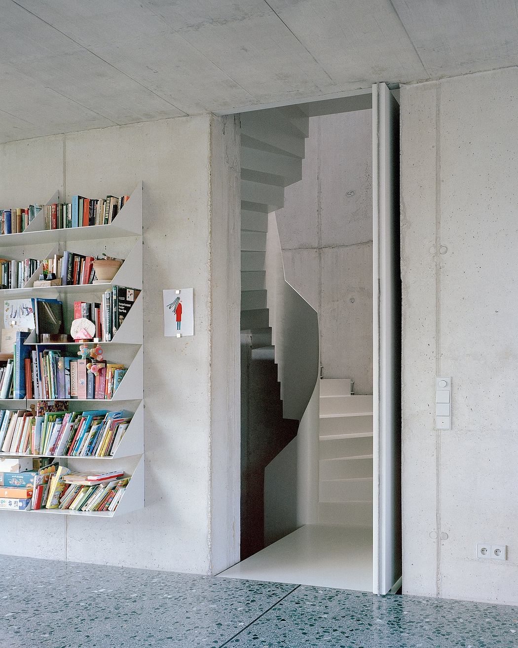 A minimalist concrete interior with built-in bookshelves and a hidden stairwell.