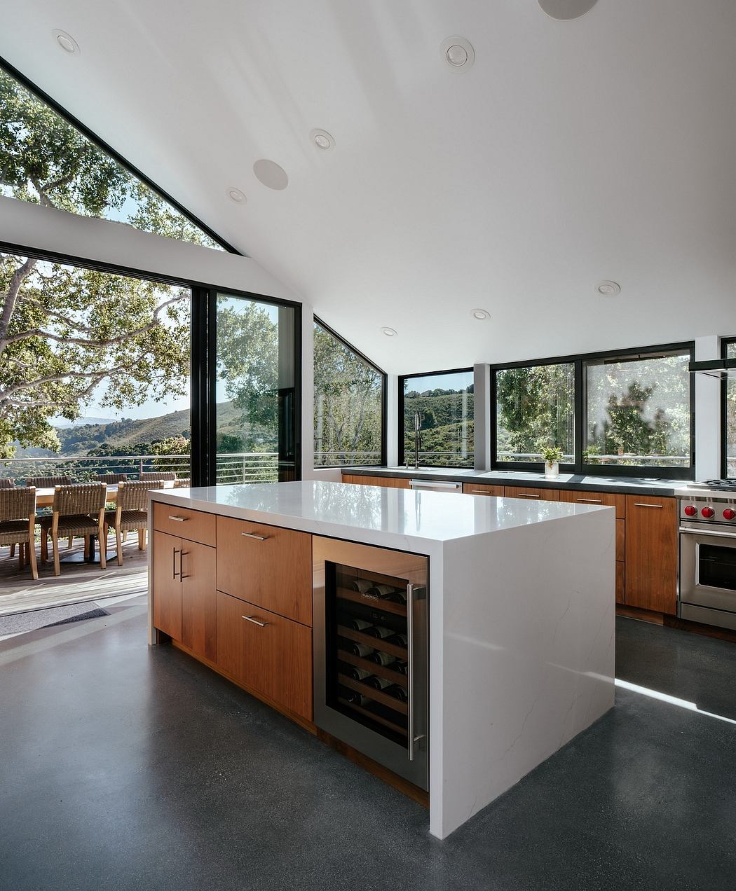 A contemporary kitchen with a sleek white island, wooden cabinets, and floor-to-ceiling windows showcasing the surrounding nature.
