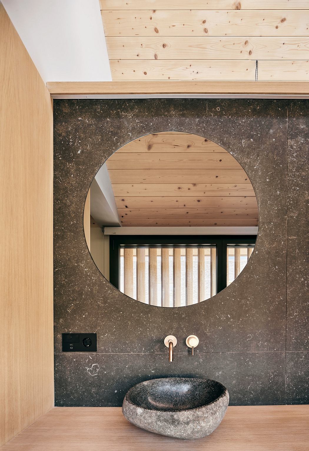 Circular mirror framed by wood-paneled ceiling, slate vanity with brass faucets.