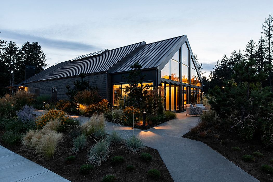 Modern A-frame house with glass walls and dark metal roof amid a lush landscaped garden at dusk.