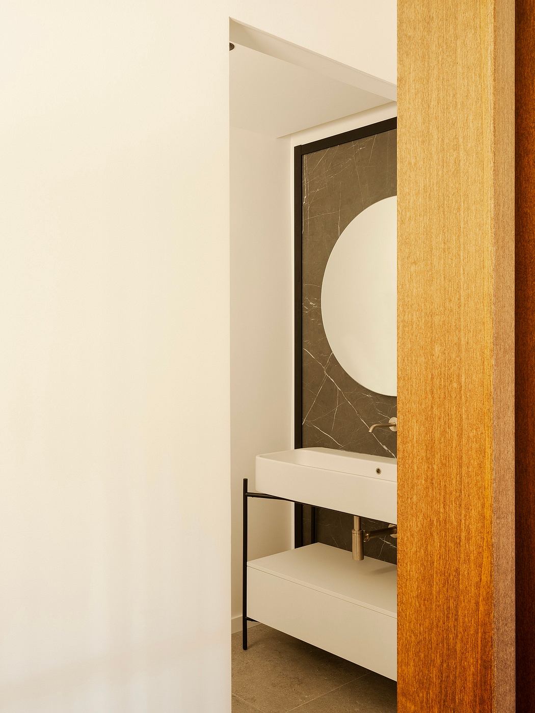 A modern bathroom with a circular mirror and a white vanity, framed by warm wooden accents.