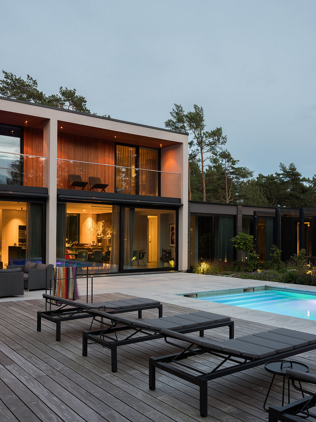 Sleek, modern home design with glass walls, poolside lounge chairs, and wooded surroundings.