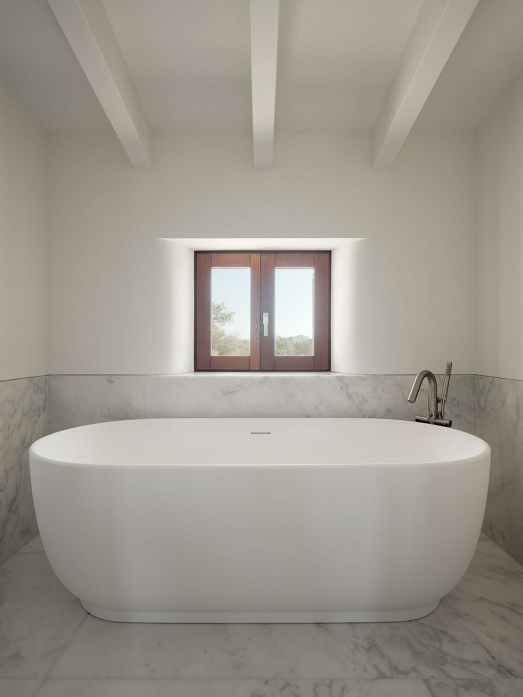 A modern bathroom with a freestanding white bathtub, marble walls, and large windows overlooking nature.