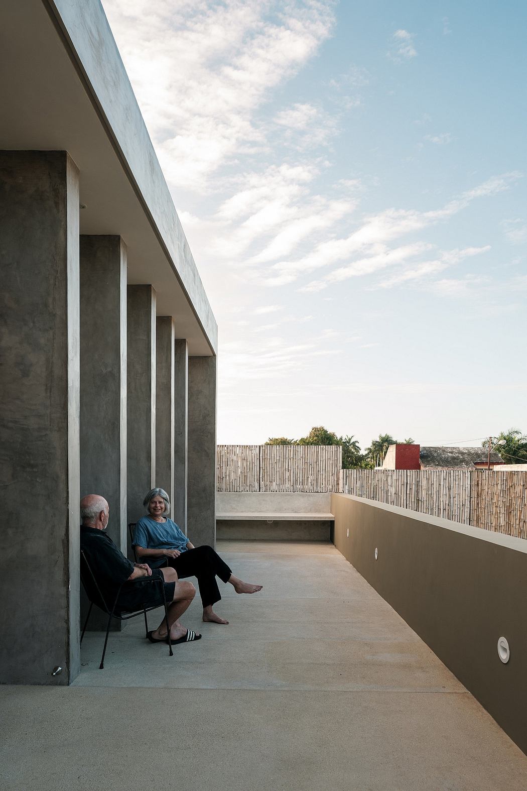 A minimalist concrete structure with bamboo fencing, and two people relaxing on a balcony.