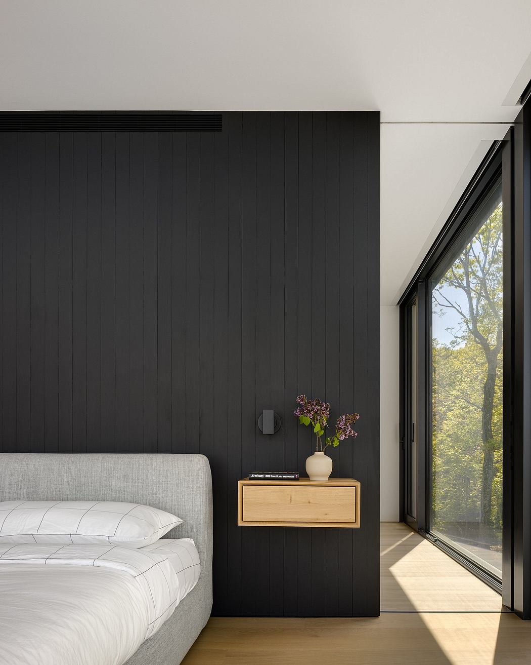 Minimalist bedroom with dark paneled walls, wooden nightstand, and large window overlooking nature.