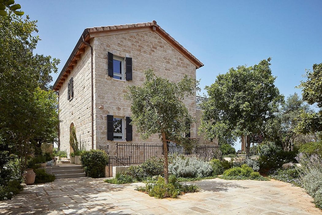 A two-story stone building with a tiled roof, surrounded by lush greenery and a paved courtyard.