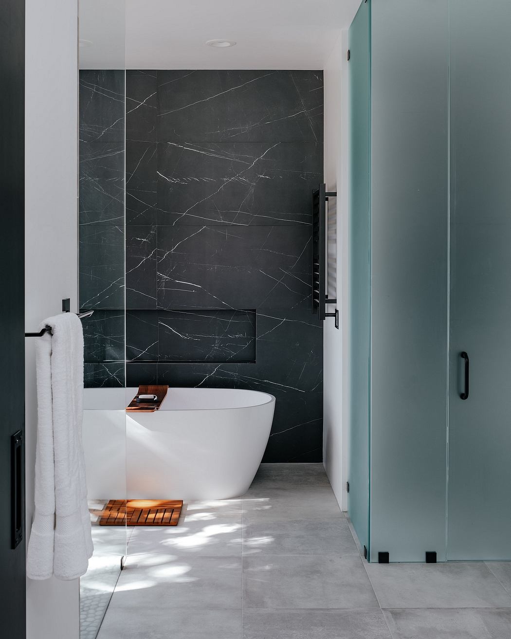 Sleek modern bathroom with black marble walls, freestanding tub, and glass partition.