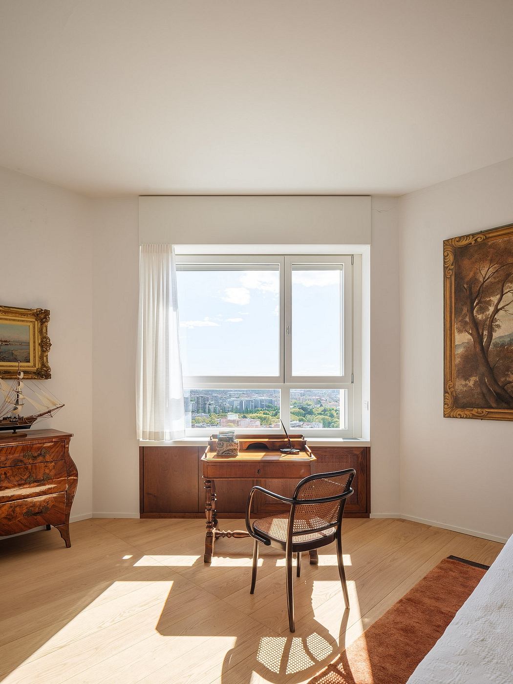 Spacious room with large window overlooking city, antique furniture, and framed artwork.