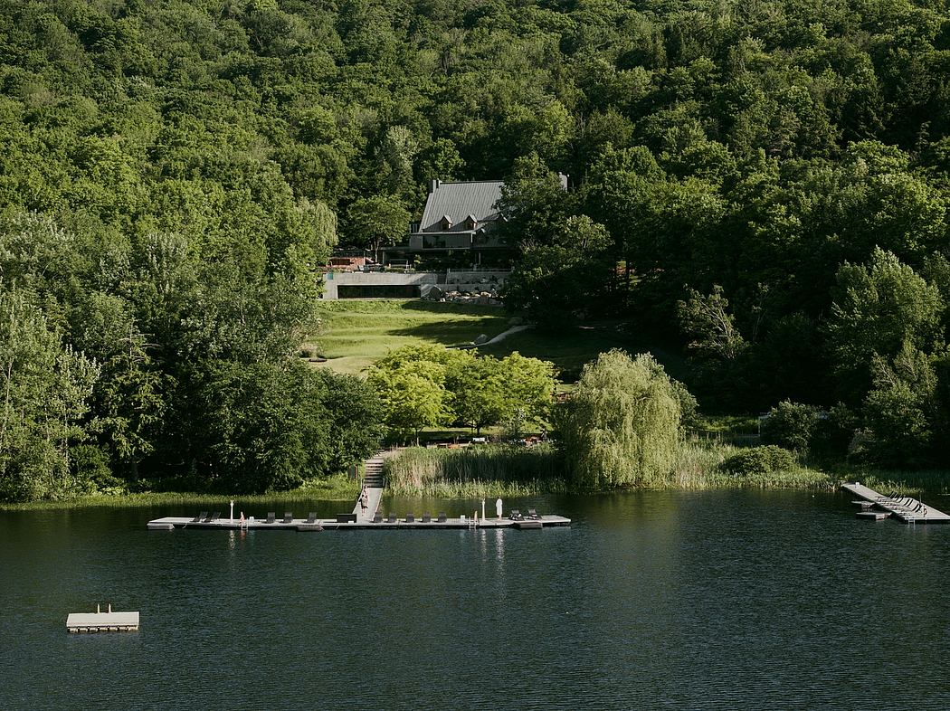 A lakefront house nestled amid lush, verdant foliage, with a wooden dock and floating platform extending into the serene waters.