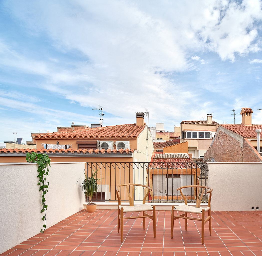 Terracotta-tiled rooftops, balcony with wood chairs, lush potted plants, and a sunny sky.