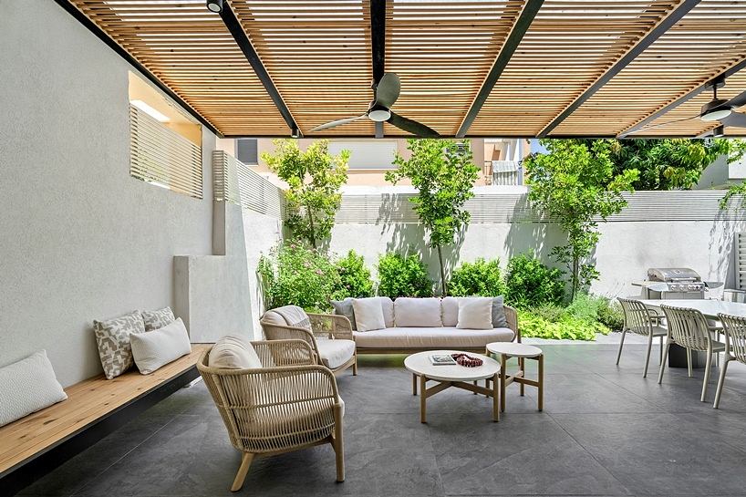 A modern outdoor living space with a slatted wooden canopy, comfortable seating, and lush greenery.