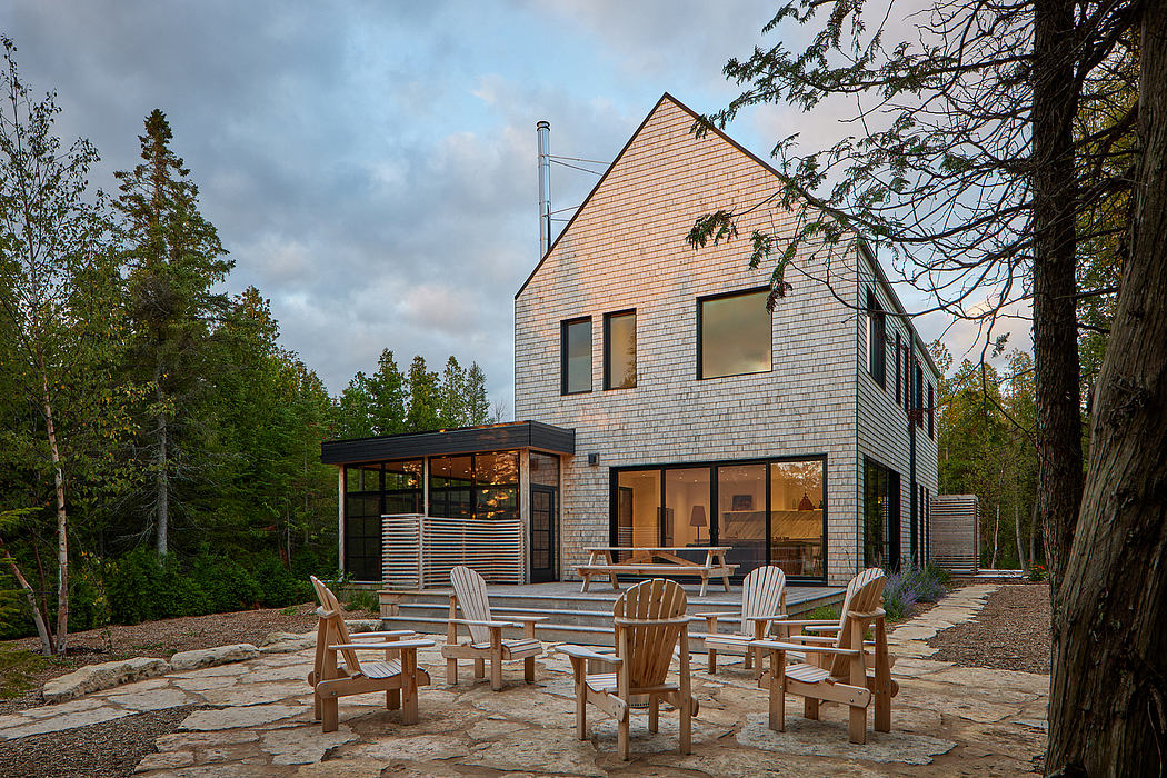 A contemporary wooden house with a covered patio and Adirondack chairs on a stone patio, surrounded by trees.