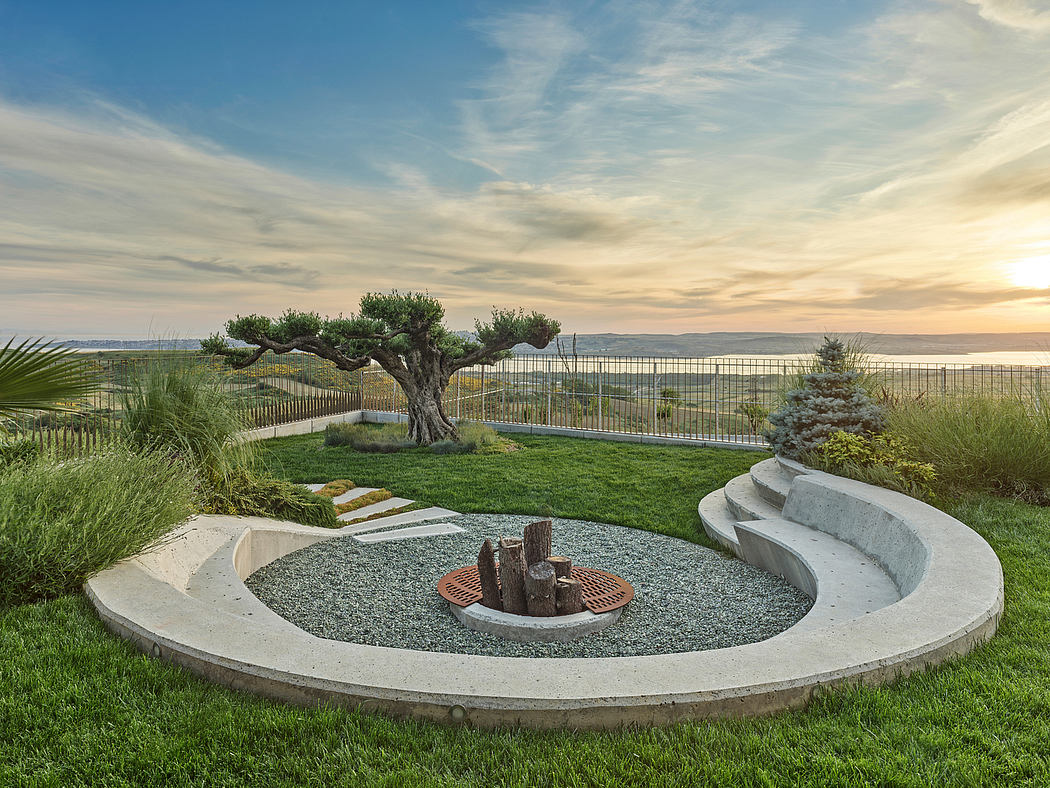 Landscaped backyard with concrete seating, fire pit, and lush greenery overlooking water.