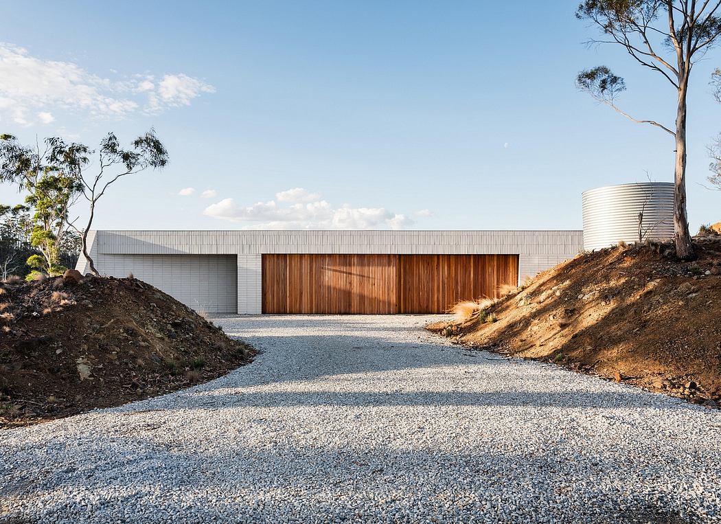 A modern, minimalist building with a large wooden door and a gravel driveway surrounded by trees.