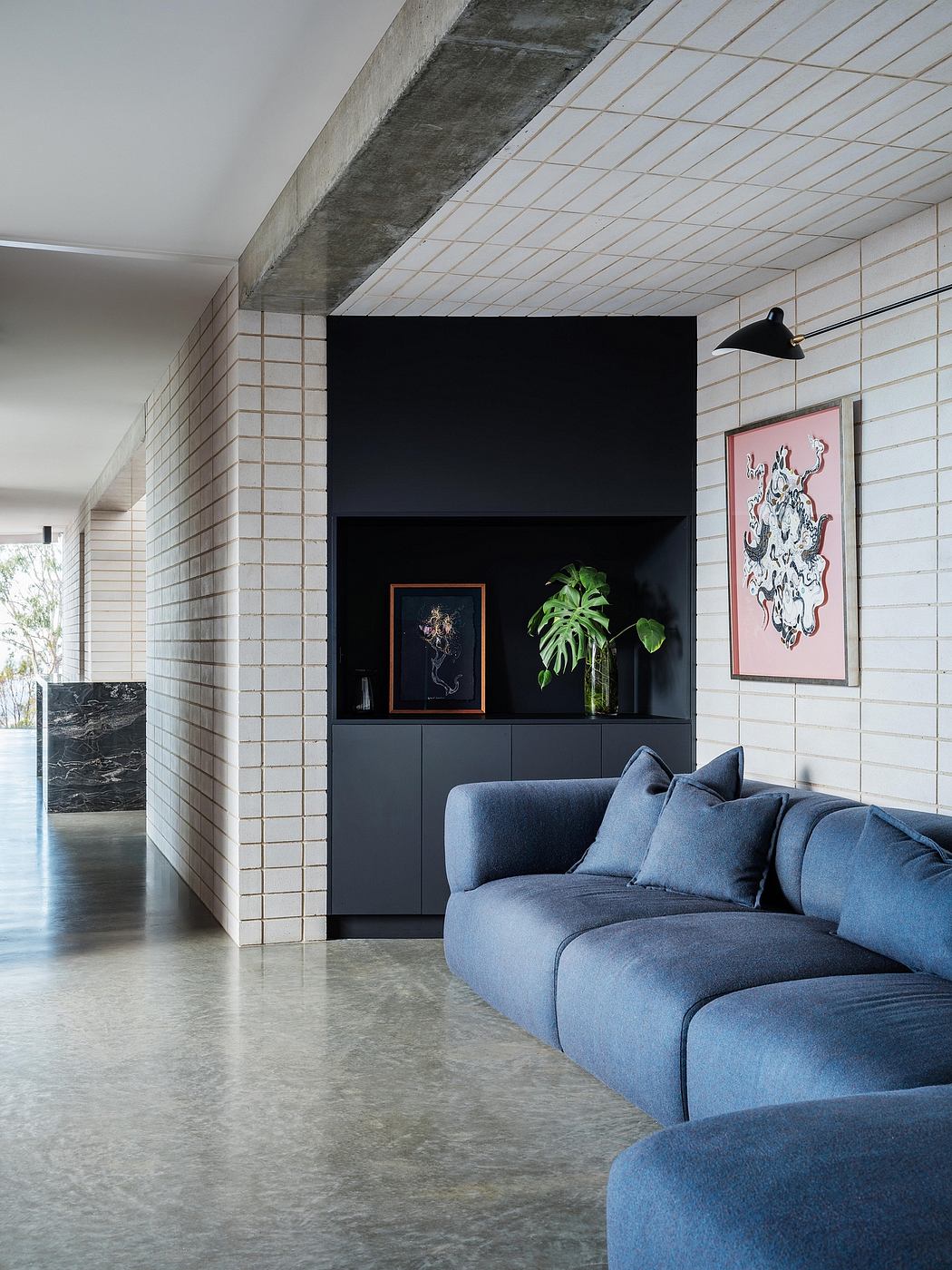 Modern living room with gray concrete floor, white brick walls, and a black wall unit displaying artwork and plants.