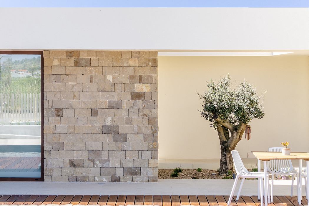 Minimalist architectural design with stone wall, olive tree, and outdoor dining area.