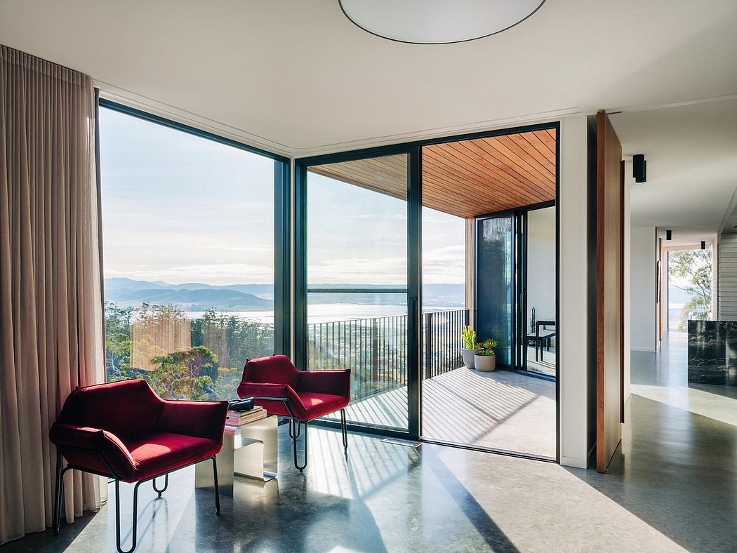 Expansive panoramic windows offer a serene waterfront view, complemented by plush red chairs.