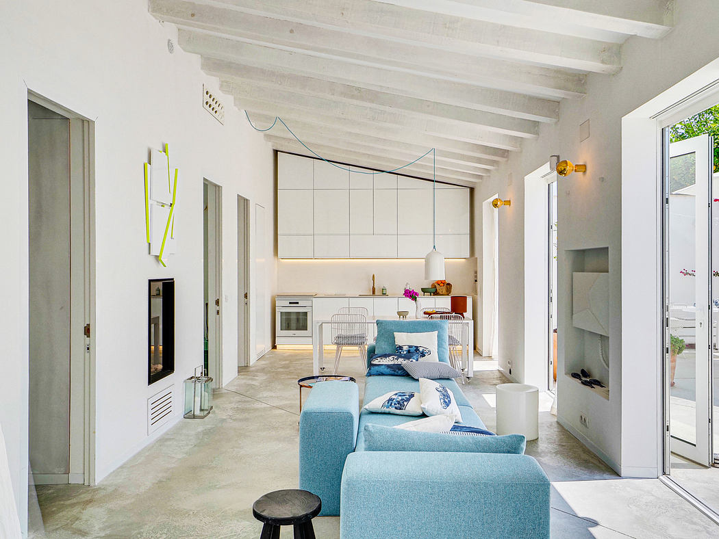 The image shows a bright, open-plan living area with wood-beamed ceilings, minimalist furnishings, and a modern kitchen.