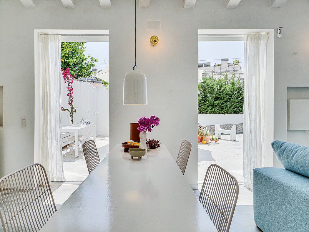 Minimalist white interior with large windows, wooden dining table, and lush outdoor greenery.