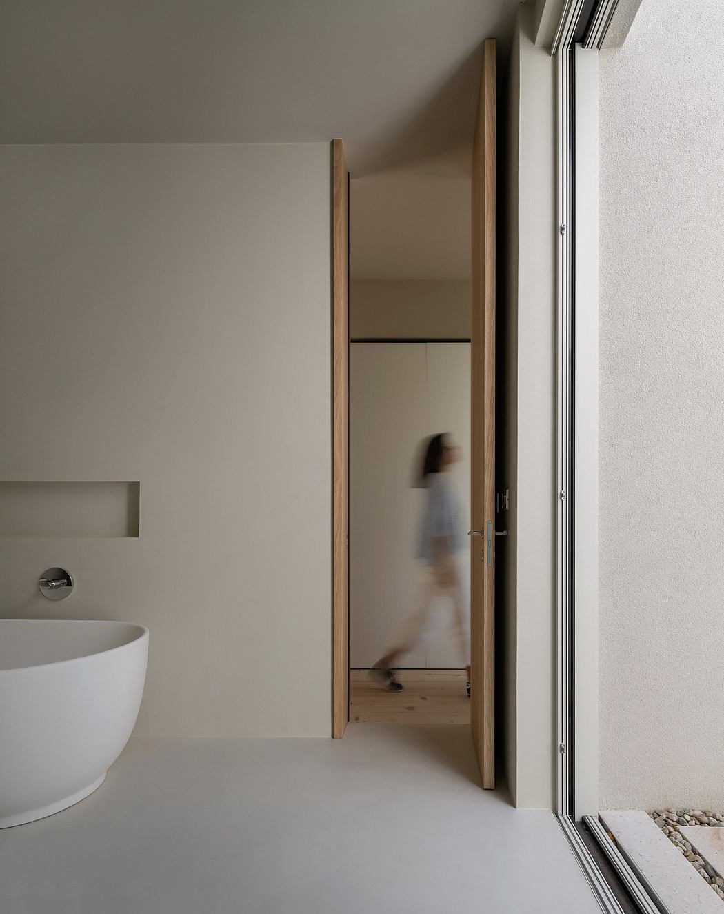 Minimalist bathroom with freestanding tub, wooden doorframe, and blurred figure moving through the frame.