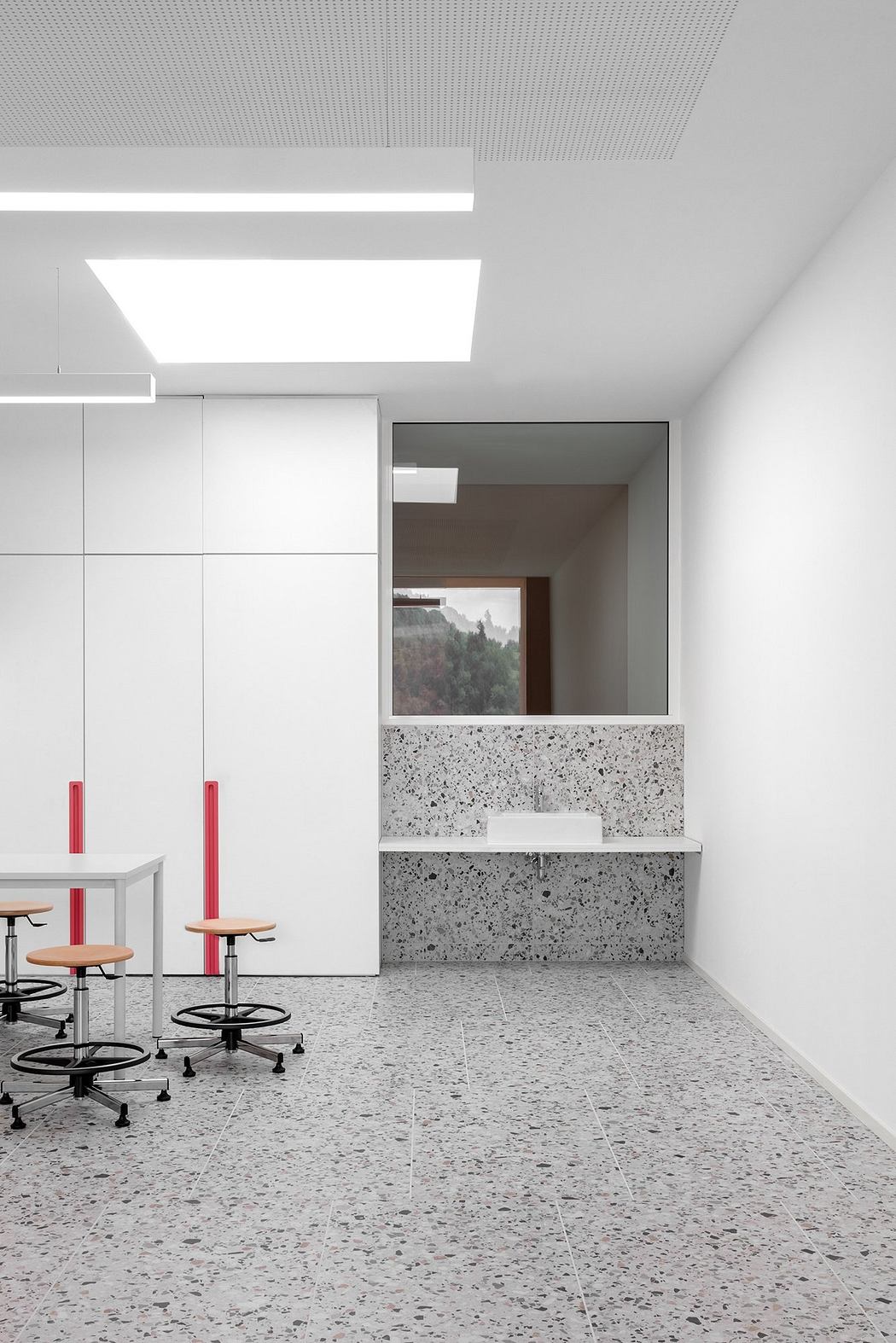 A modern, minimalist office space with terrazzo flooring, white walls, and red accents.