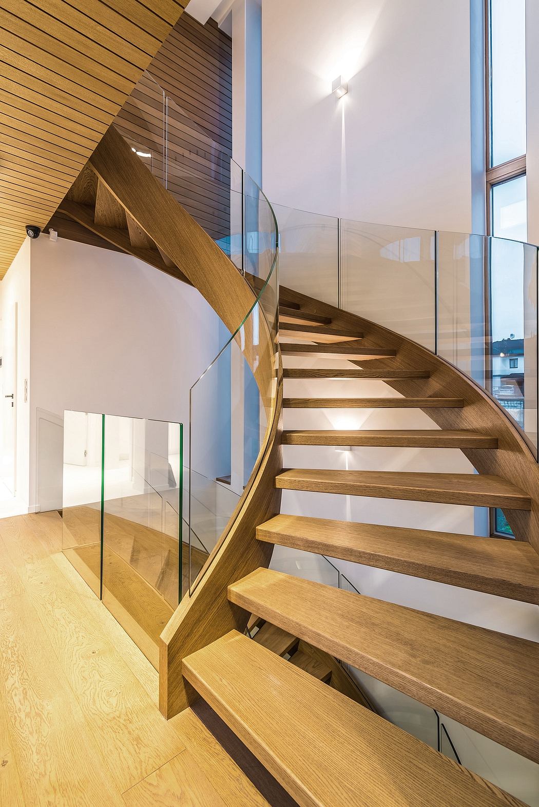 A modern, open staircase with curved wooden steps and glass railings, accentuated by natural lighting.