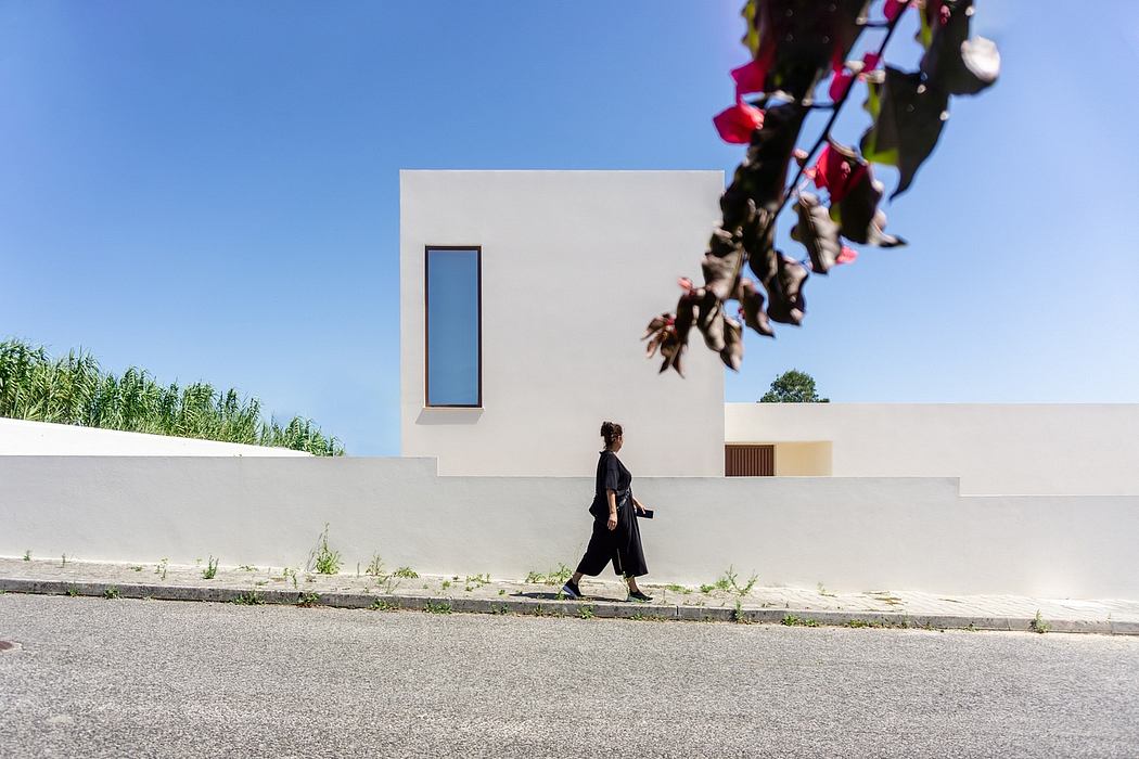 A modern, minimalist building with a single narrow window and vegetation surrounding it. A person in black clothing walking by.