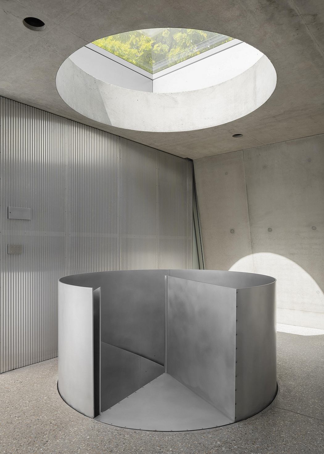 Circular skylight illuminates minimalist concrete interior with abstract metal pedestal.
