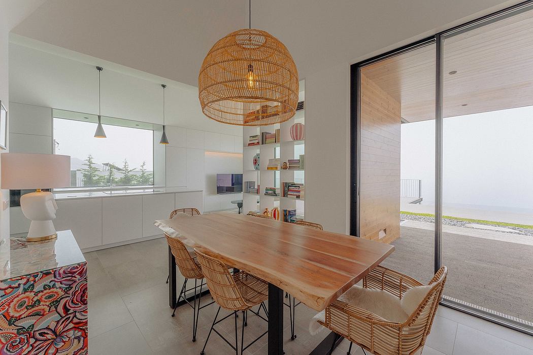 A modern, light-filled kitchen with a wooden dining table, woven rattan pendant lamps, and shelving displaying decorative items.