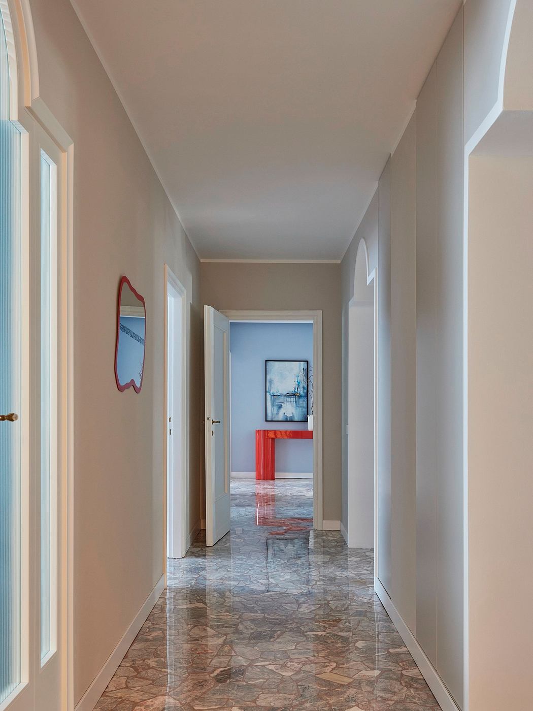 Hallway with beige walls, marble floors, and red accent table framing artwork.